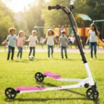 קורקינט ילדים שלושה גלגלים בצבע ורוד על דשא, עם ילדים מטושטשים ברקע בפארק | yrz