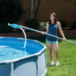 אישה מנקה בריכה עילית עם מוט ושואב/רשת ניקוי המחוברים לבריכה | yrz