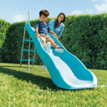 ילד גולש במגלשה כחולה בחצר עם סולם מתכת, כשמבוגר משגיח מאחור | yrz