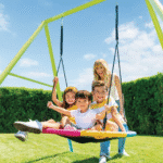 שלושה ילדים יושבים על נדנדת צלחת צבעונית בחצר, עם מבוגר שעומד מאחור | yrz