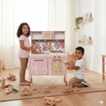 שני ילדים משחקים במטבח עץ לילדים בצבע ורוד בסלון עם צעצועי עץ על הרצפה